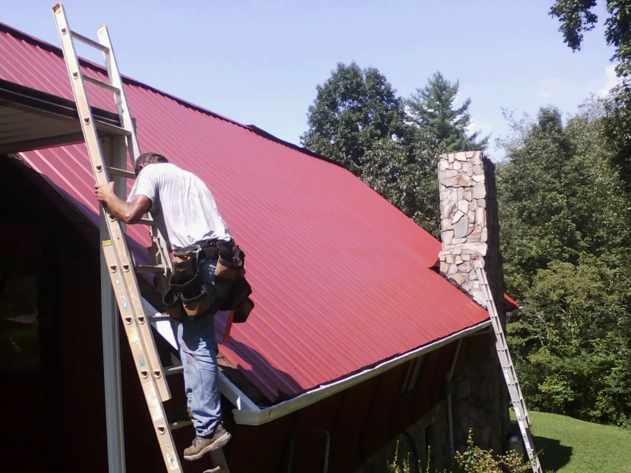 Roofer on a ladder beside a red metal roof and stone chimney — Soffit & Fascia Repair by Roofing Brothers in Kenilworth